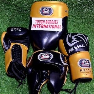 May include: A collection of boxing equipment, including a black and gold punching bag, sparring gloves, and a head guard. The gloves are labeled "RS11V SPARRING GLOVES 14 OZ". The text "TOUGH BUDDIES INTERNATIONAL" is visible on a white sign.