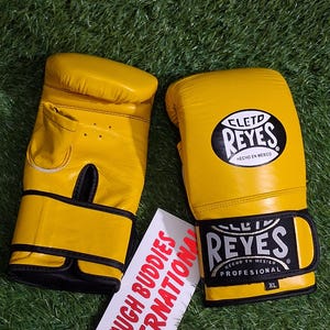 May include: A pair of yellow boxing gloves with black accents and the brand name "Reyes" visible. The gloves are on a green surface. A white tag with the text "TOUGH BUDDIES INTERNATIONAL" is also present.