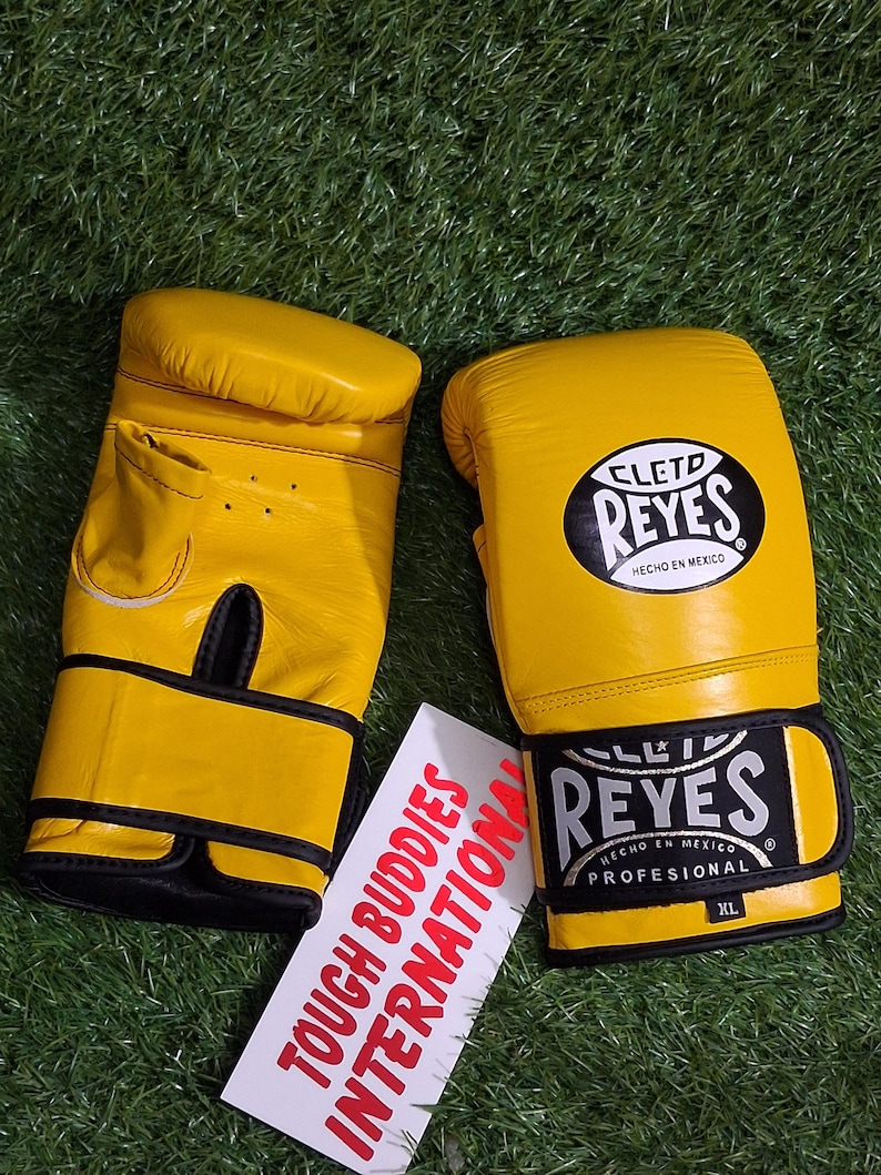 May include: A pair of yellow boxing gloves with black accents and the brand name "Reyes" visible. The gloves are on a green surface. A white tag with the text "TOUGH BUDDIES INTERNATIONAL" is also present.