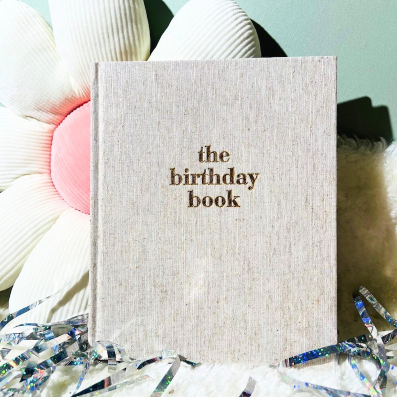 May include: A beige linen-textured book titled "the birthday book" in gold lettering. The book is propped up against a large white and pink flower-shaped pillow. Silver and iridescent confetti is scattered around the book.