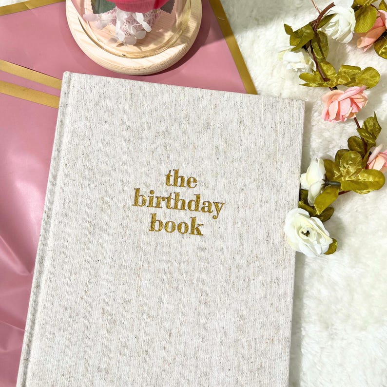 May include: A hardcover book with a textured, light gray cover. The gold text reads "the birthday book." The book is surrounded by pink and white flowers and sits on a pink and gold surface.