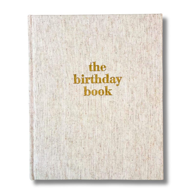 May include: A hardcover book with a beige linen cover. The words "the birthday book" are printed in gold lettering on the front. The book is rectangular and has a clean, minimalist design.