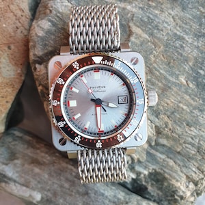 May include: A silver wristwatch with a square face and a mesh strap. The watch face has a silver dial with red and white details, and the text "FIFTYFIVE Fathoms" is visible. The bezel is black and red, with white numerals.