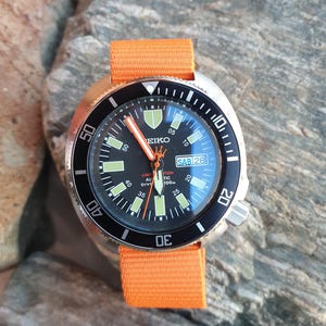Op de afbeelding: Een Seiko duikhorloge van roestvrij staal met een zwarte lunette en wijzerplaat, lichtgevende markeringen en wijzers. Oranje nylon band. De wijzerplaat toont de dag en datum. Het horloge ligt op een getextureerd grijs oppervlak.