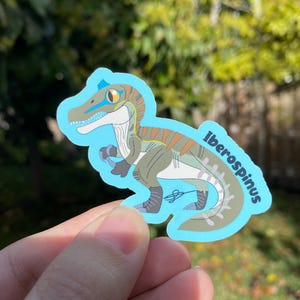 May include: A sticker featuring a cartoon illustration of an Iberospinus dinosaur in shades of green, brown, and blue. The dinosaur has a blue stripe across its eye and the name "Iberospinus" is written on the sticker.