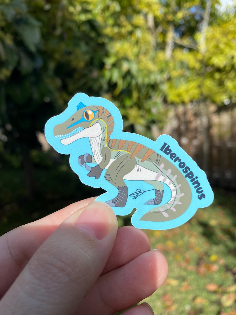 May include: A sticker featuring a cartoon illustration of an Iberospinus dinosaur in shades of green, brown, and blue. The dinosaur has a blue stripe across its eye and the name "Iberospinus" is written on the sticker.