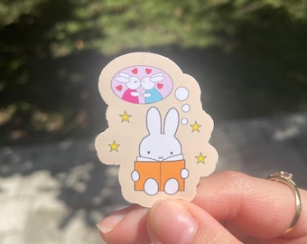 May include: A sticker featuring a white rabbit reading an orange book, with a thought bubble showing two rabbits in love. The sticker has a beige background with yellow stars and a pink and blue heart design.