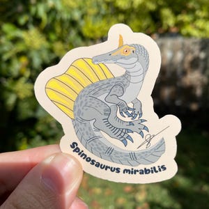 May include: A sticker featuring a Spinosaurus mirabilis dinosaur illustration. The dinosaur is depicted in shades of gray with a yellow sail and blue claws. The sticker has a beige border and the text "Spinosaurus mirabilis" is printed on it.