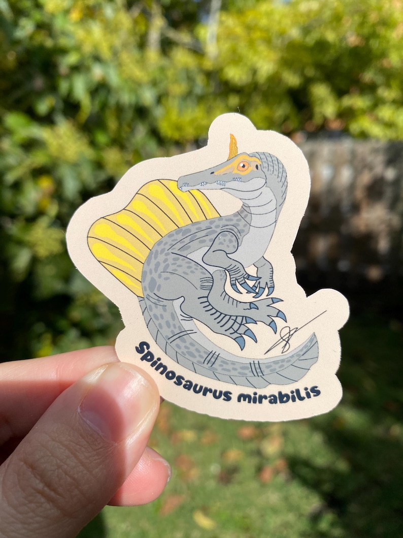 May include: A sticker featuring a Spinosaurus mirabilis dinosaur illustration. The dinosaur is depicted in shades of gray with a yellow sail and blue claws. The sticker has a beige border and the text "Spinosaurus mirabilis" is printed on it.
