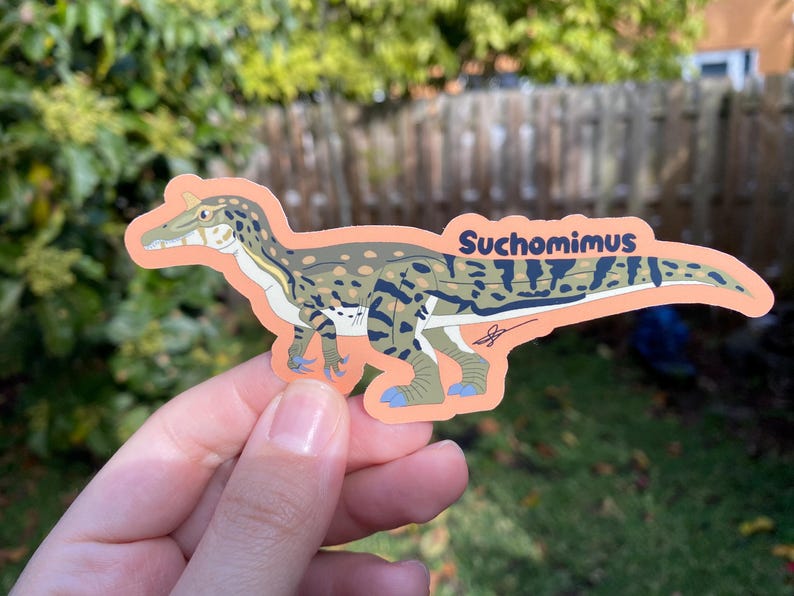 May include: A Suchomimus sticker featuring a detailed illustration of the dinosaur in shades of green, brown, and blue, with the name "Suchomimus" printed in blue. The sticker has an orange border and is held by a hand.