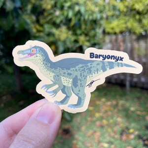 May include: A sticker featuring a Baryonyx dinosaur illustration. The dinosaur is primarily blue and green with a tan background. The word "Baryonyx" is written in a dark blue font. The sticker is held by a person's hand.