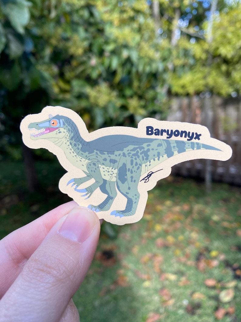 May include: A sticker featuring a Baryonyx dinosaur illustration. The dinosaur is primarily blue and green with a tan background. The word "Baryonyx" is written in a dark blue font. The sticker is held by a person's hand.