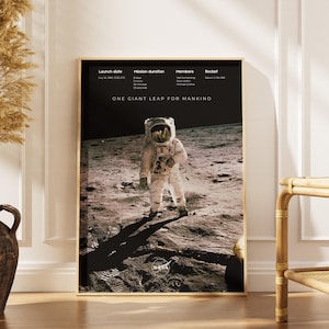 Puede incluir: Un póster enmarcado con un astronauta en la luna y el texto "ONE GIANT LEAP FOR MANKIND". La imagen está en tonos sepia, mostrando la sombra del astronauta en la superficie lunar. El póster se exhibe sobre un suelo de madera.
