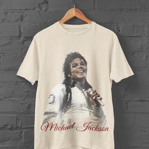 May include: Off-white t-shirt featuring a smiling image of Michael Jackson holding a microphone. The text "Michael Jackson" is written in a cursive font below the image. The shirt is hanging on a wooden hanger.