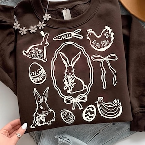 May include: A dark brown sweatshirt featuring white Easter-themed graphics, including rabbits, eggs, carrots, and a chicken. The sweatshirt is paired with blue jeans and a floral necklace. A brown hat is visible in the background.