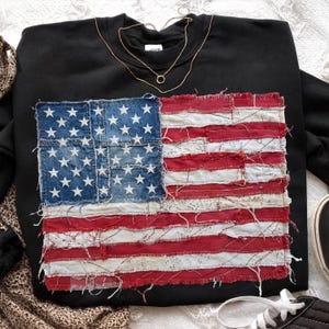 Könnte beinhalten: Schwarzes Sweatshirt mit einem Patchwork-Design der amerikanischen Flagge. Die Flagge besteht aus Denim und rot-weißem Stoff. Eine goldene Halskette ist auf dem Sweatshirt drapiert. Das Sweatshirt befindet sich auf einer weißen Oberfläche.