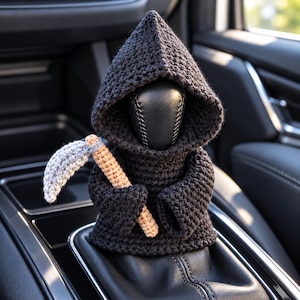 May include: A black crocheted figure wearing a hooded cloak, designed to fit over a car gear shift knob. The figure holds a small, light brown and grey crocheted scythe in its hands, featuring a textured yarn construction throughout the piece.
