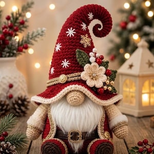 Puede incluir: Una figura de gnomo de ganchillo con un sombrero rojo alto y rizado decorado con copos de nieve blancos, una flor beige, hojas verdes y bayas rojas. El gnomo tiene una cara beige, una larga barba blanca, un cinturón marrón y botas de color rojo oscuro.