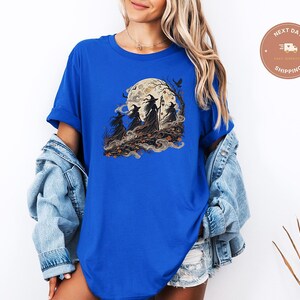 May include: A royal blue short-sleeved t-shirt featuring a graphic print of three witches in pointed hats walking through a forest under a large full moon. The design includes swirling mist, autumn leaves, and a bird silhouette against the moon.