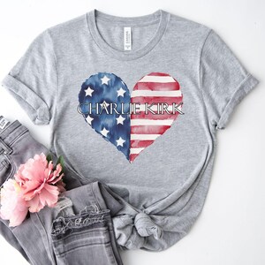 A gray t-shirt with a heart-shaped design featuring the text "CHARLIE KIRK" and an American flag pattern, along with a pink flower.