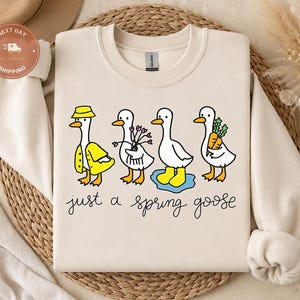 May include: A beige crewneck sweatshirt featuring a graphic of four white geese. One goose wears a yellow raincoat and hat, one holds flowers, one stands in a blue puddle wearing yellow boots, and one holds carrots. The text reads just a spring goose.