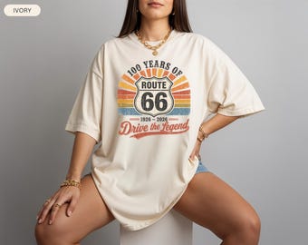 May include: Ivory-colored t-shirt featuring a Route 66 graphic. The design includes the text "100 Years of Route 66" and "Drive the Legend" in a vintage style, with the years 1926-2026. The shirt has a relaxed fit.