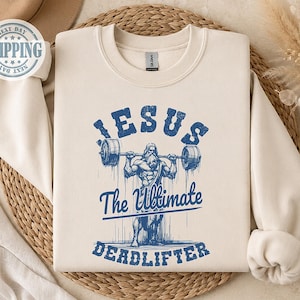 May include: A cream-colored crewneck sweatshirt featuring a blue graphic print of a figure lifting a barbell. The text reads "JESUS The Ultimate DEADLIFTER" in a distressed font. The design includes vertical lines extending downward from the figure.