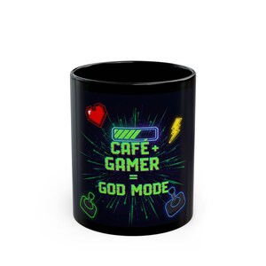 Puede incluir: Taza de café negra con el texto "CAFE + GAMER = GOD MODE" en verde neón. La taza presenta un corazón de píxeles rojo, un rayo y iconos de mando de juego. El diseño está sobre un fondo oscuro.
