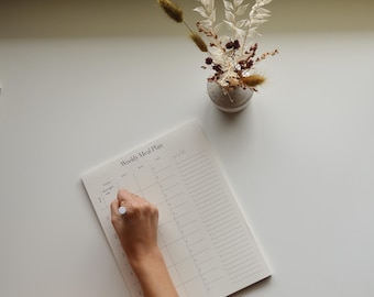 Op de afbeelding: Een hand schrijft met een pen op een notitieblok met de tekst "Weekly Meal Plan". Het notitieblok ligt op een wit oppervlak, naast een kleine vaas met gedroogde bloemen. Het notitieblok heeft kolommen voor elke dag van de week.