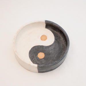 Puede incluir: Un plato redondo de cerámica con un diseño de yin y yang. El plato está dividido en dos secciones: una blanca y otra negra. Dos pequeños acentos redondos de color dorado están en el centro de cada sección. El plato está sobre un fondo blanco.