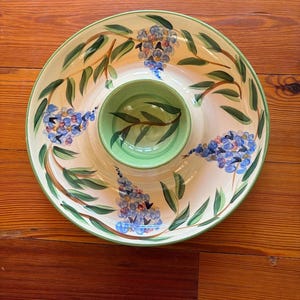 May include: A round ceramic serving dish with a central dip bowl, featuring a cream-colored base decorated with hand-painted blue flower clusters and green leaves. The rim and the interior of the center bowl are finished in a light green glaze.