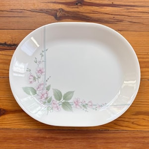 May include: White oval serving platter with a floral design. The pattern features pink flowers, green leaves, and subtle gray and blue lines. The platter is set against a wooden background.