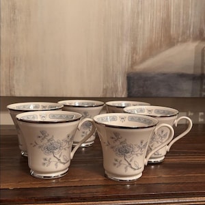 May include: Set of six cream-colored teacups with a floral design in blue and silver trim. Each cup features a delicate floral pattern and a silver rim. The cups are arranged on a wooden surface.