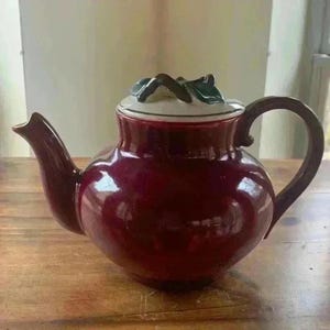 May include: A burgundy ceramic teapot with a rounded body, a curved spout, and a dark brown handle. The lid is white with a green and brown leaf design. The teapot is sitting on a wooden surface.
