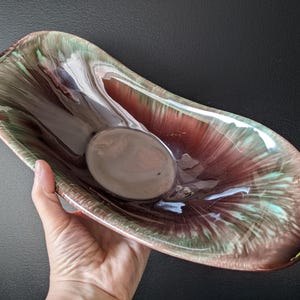 Può includere: Un piatto vintage in ceramica oblunga con una forma unica e una finitura lucida. Il piatto presenta una miscela di colori, tra cui marrone, verde e un pizzico di blu. Il piatto ha una piccola area incassata rotonda al centro. Il piatto è tenuto da una mano.