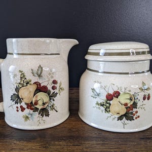 Può includere: Set di brocca e barattolo in ceramica color crema. Entrambi i pezzi presentano un disegno di frutta con pere, ciliegie e uva. La brocca ha un manico e un beccuccio, mentre il barattolo ha un coperchio. Accenti verdi e marroni completano il design.