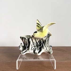 Puede incluir: Una maceta de cerámica con forma de tocón de árbol con acabado brillante, que presenta una flor esculpida en la parte frontal y un pequeño pájaro amarillo y verde posado en el borde con las alas parcialmente extendidas, en tonos gris y blanco.