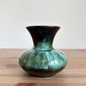 May include: A small ceramic vase with a rounded body and flared rim. The vase has a glossy glaze in shades of green and brown, creating a marbled effect. The vase is displayed on a wooden surface.