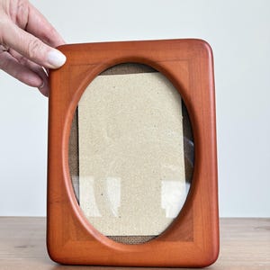 Könnte beinhalten: Ein rechteckiger Bilderrahmen aus Holz in warmem Rotbraun mit ovaler Öffnung. Der Rahmen hat eine Glasfront und eine Kartonrückseite. Der Rahmen wird von einer Hand gehalten.