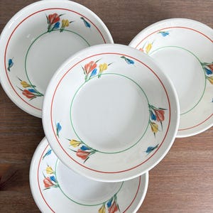 Pode incluir: Quatro tigelas de cerâmica branca com um design floral. Cada tigela tem uma borda vermelha, um círculo interno verde e detalhes florais e de folhas coloridos em vermelho, amarelo e azul. As tigelas estão empilhadas em uma superfície de madeira.