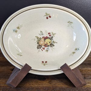 May include: Cream-coloured oval serving platter with a fruit design. The platter has a brown rim and a central fruit arrangement of pears, berries, and apples. It is supported by a wooden stand.