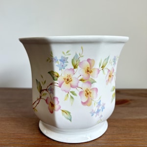 May include: A white ceramic planter with an octagonal shape and a flared rim. The exterior features a delicate floral pattern with pink and yellow blossoms, green leaves, and small blue flowers arranged in a trailing vine design around the body.