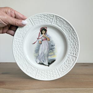 May include: A white ceramic plate featuring a central illustration of a person in a long white dress and pink bonnet, with the text Pinkie Lawrence. The rim has a raised basket weave and floral pattern around the circular edge of the plate.