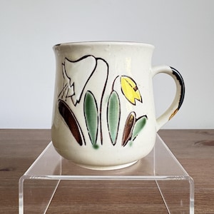 May include: A cream-coloured ceramic mug featuring a hand-painted floral design with a large white bell-shaped flower, a smaller yellow bud, and green and brown leaves. The mug has a curved handle with a dark brown accent at the top.