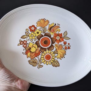 May include: Oval white serving platter with a floral design. The flowers are in shades of orange, yellow, and brown. The platter has a ridged border and is held by a hand.