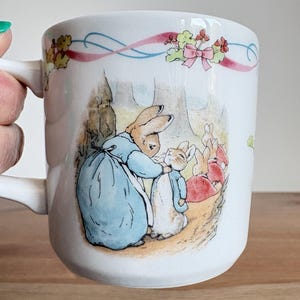 Peut inclure: Mug en céramique blanche avec une illustration classique de lapins dans une forêt. La tasse a une anse et est décorée d'un motif de ruban rose et bleu. L'illustration représente une mère lapin et ses enfants.