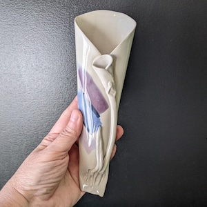 Op de afbeelding: Een crèmekleurige keramische wandvaas in een unieke, gevouwen vorm. De vaas heeft een verhoogd calla lelie-ontwerp en abstracte paarse en blauwe geschilderde accenten. De vaas wordt door een hand tegen een donkere achtergrond gehouden.