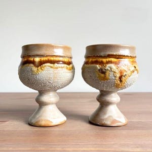 May include: Two ceramic goblets featuring a textured, speckled off-white base and a smooth, glossy rim with a drip-like glaze in shades of amber, brown, and cream. Each vessel has a wide bowl, a thick stem, and a flared circular foot base.