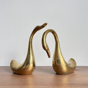 May include: Two brass swan figurines. The swans have long, curved necks and are positioned facing each other. The figurines are golden in color and sit on a wooden surface. The background is a plain white wall.
