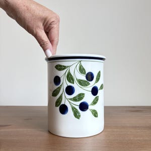 Puede incluir: Un recipiente de cocina cilíndrico de cerámica blanca con un patrón decorativo de bayas azul oscuro y hojas verdes. El contenedor tiene un acabado liso con un borde azul oscuro en la parte superior, ofreciendo un recipiente de almacenamiento funcional.